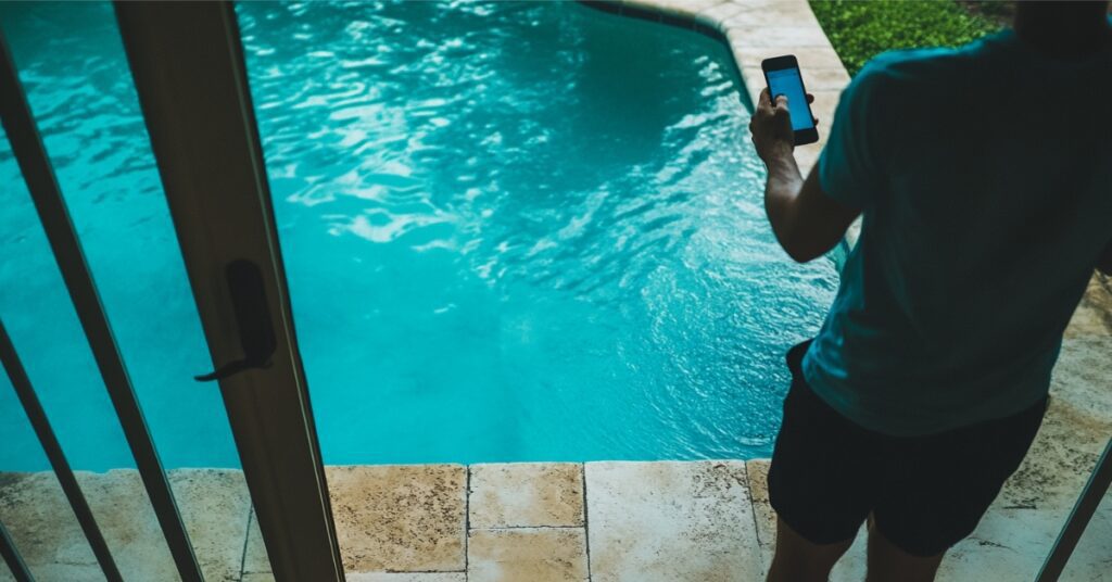 hand holding cell phone over pool, concept of Why Smart Pools Are the Future: Guide for Beaumont Residents
